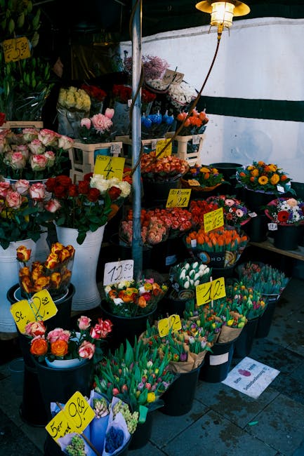 A colorful outdoor flower stall displays a variety of fresh flowers and floral arrangements in black buckets and baskets. There are pink and white roses, red roses, and mixed flower bouquets with brig