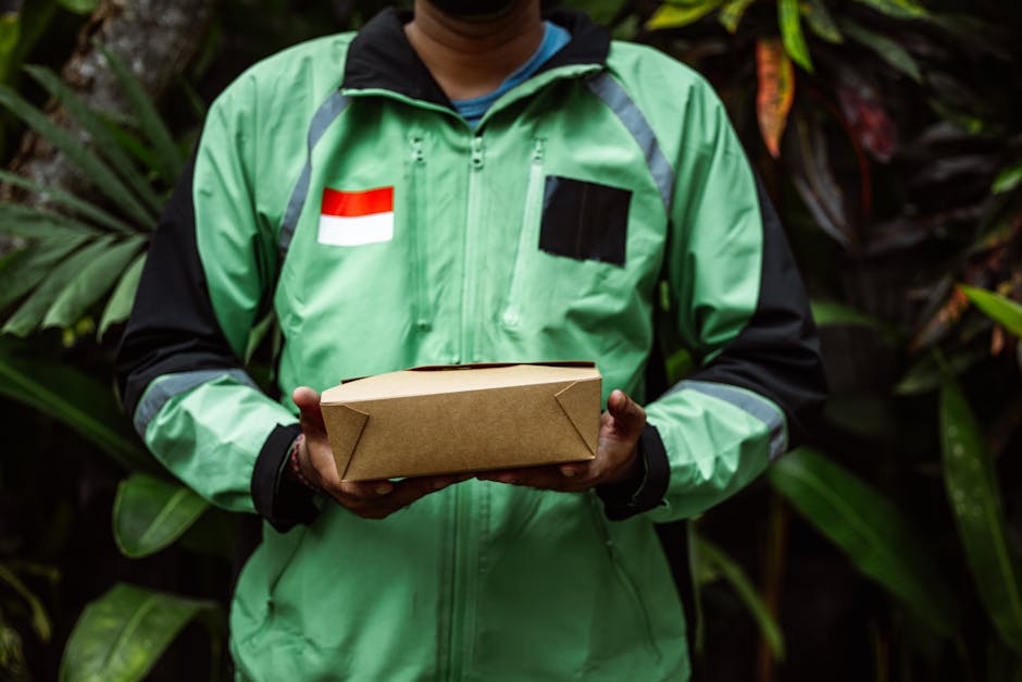 A person wearing a green and black jacket with a red and white flag patch on the left chest, holding a small rectangular cardboard box with both hands. The individual is standing outdoors amidst lush