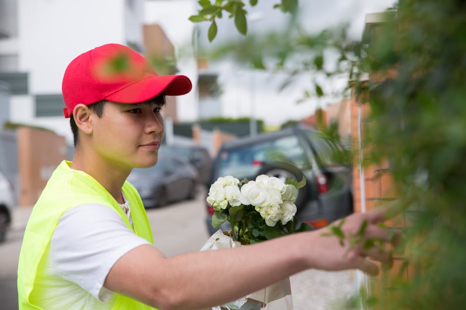 A young flower delivery person wearing a red cap and a yellow safety vest is arranging a bouquet of fresh white roses and green foliage against a brick wall outdoors. The bouquet is wrapped in white p