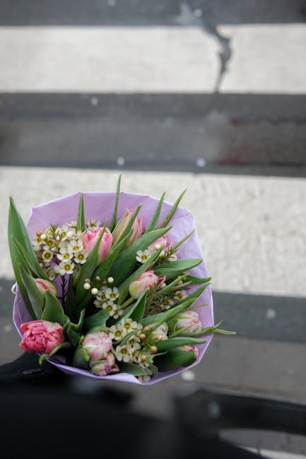 A close-up view of a floral bouquet featuring pink tulips, small white daisies, and sprigs of tiny white filler flowers, arranged with long green leaves. The bouquet is wrapped in soft lavender tissue