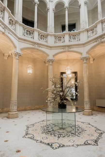 The image features an elegant, spacious interior with a high, curved ceiling supported by ornate classical columns. Natural light filters through tall, narrow windows, illuminating the cream-colored w