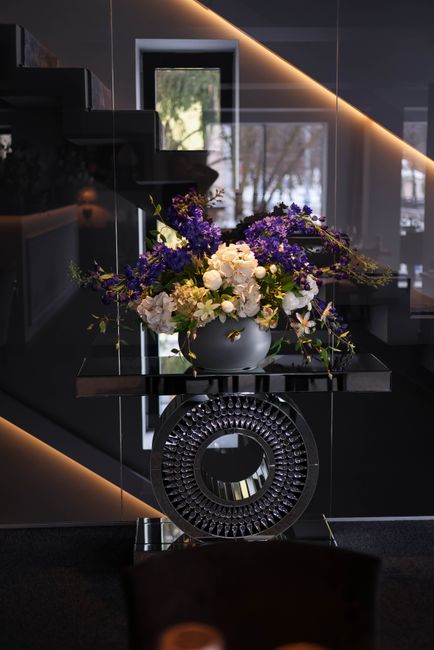A floral arrangement with a mix of white roses, purple statice, and other purple and green foliage, displayed in a gray rounded vase. The bouquet is artfully arranged with a flowing, cascading style, 