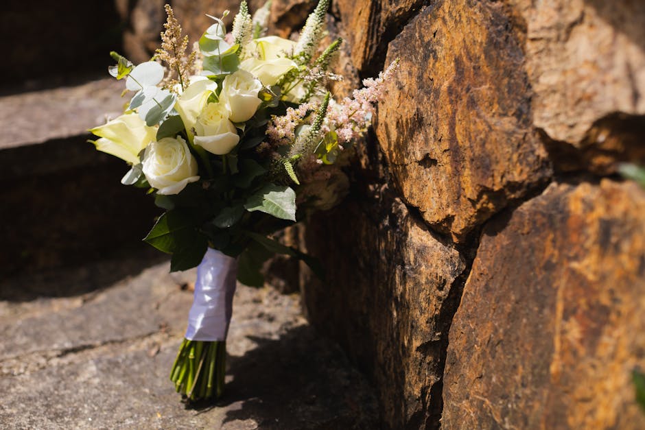 A bouquet of fresh white roses and pink astilbe flowers, arranged with green foliage and wrapped with white satin ribbon, resting against a rustic stone wall on a sunny day. The flowers display soft, 