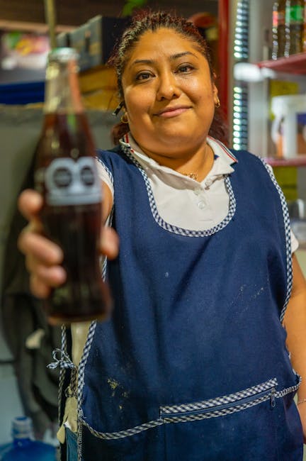 A woman holding a glass bottle of dark soda, wearing a blue apron over a white shirt, standing in a shop with shelves of colorful products in the background. The focus is on her smiling face and the b