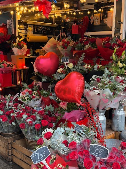 A vibrant display of fresh flowers arranged for floral gifting at a flower stall, featuring red roses, white flowers, and greenery, accented with red heart-shaped balloons. The arrangement includes bo