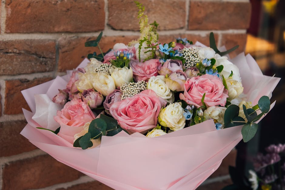 A floral bouquet arrangement featuring large, soft pink roses, white roses, and small blue forget-me-nots, accented with sprigs of white wax flowers and green eucalyptus leaves. The flowers are wrappe