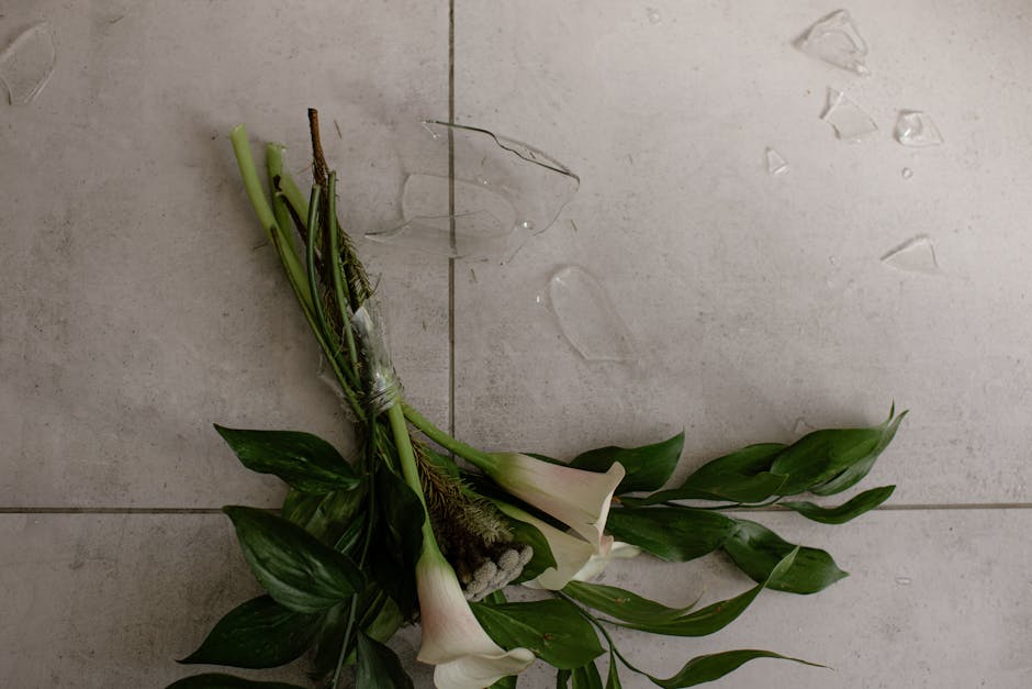 A discarded floral arrangement featuring white calla lilies with smooth, trumpet-shaped petals and elongated green leaves, lying on a light-colored tiled floor next to broken glass shards. The bouquet