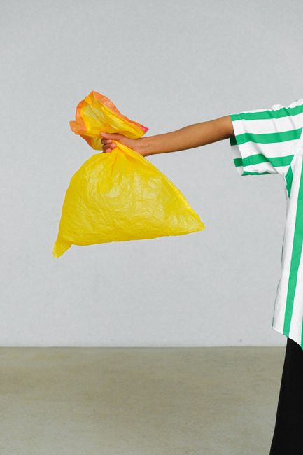 A person wearing a white and green striped shirt is holding a compostable yellow and orange paper bag filled with flower waste, with their arm extended to the right. The photo is taken against a plain