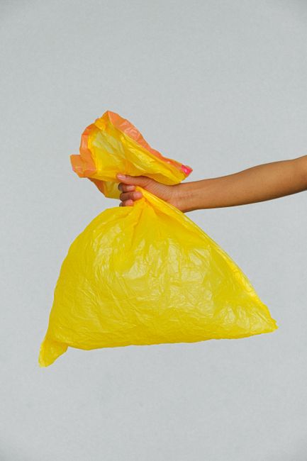 A hand holding a yellow and orange plastic bag, used for disposing of floral waste, against a plain light grey background. The bag appears to be lightweight and crinkly, with a partly visible orange t