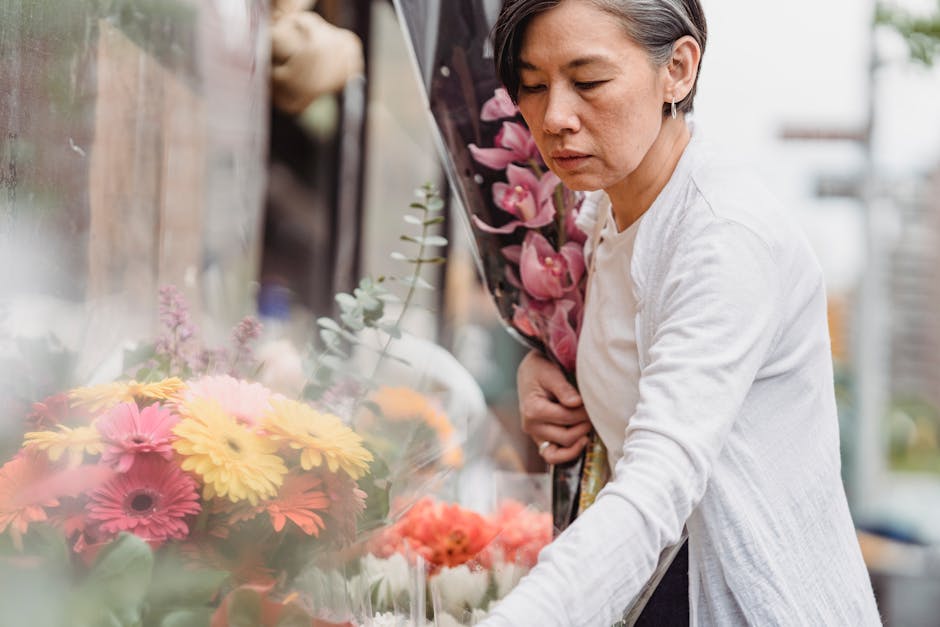 A woman with short, dark hair and a focused expression selecting a floral bouquet at a flower shop. She is holding a large arrangement of pink lilies wrapped in clear plastic in her left arm, while re