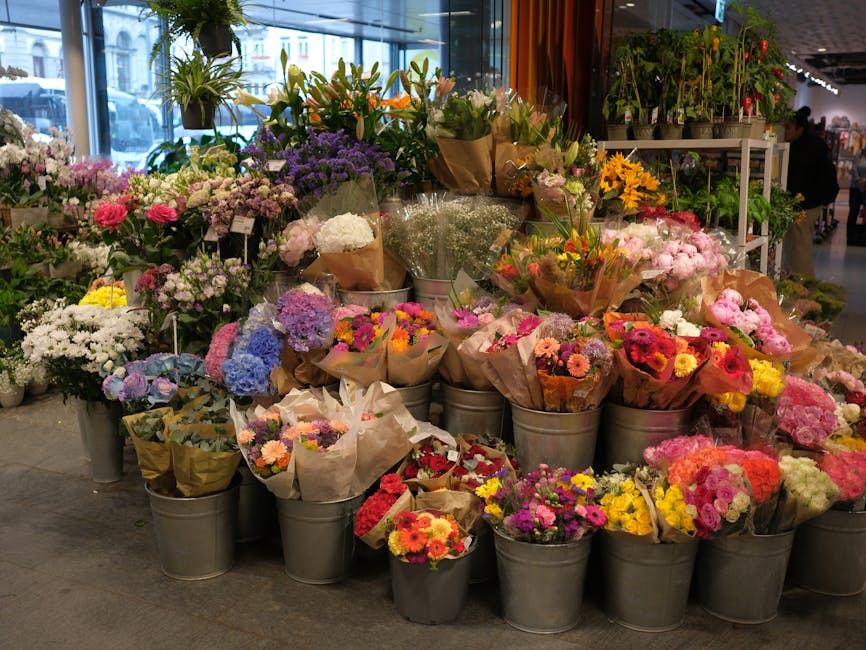 A vibrant display of fresh flower bouquets arranged in metal buckets and wrapped in brown paper, featuring a variety of blooms including pink roses, yellow daisies, purple hydrangeas, white chrysanthe